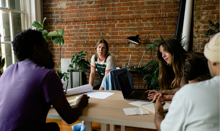 Four people sitting around a desk in an urban looking office with brick walls and plants. One person is on a laptop, while another is writing down notes